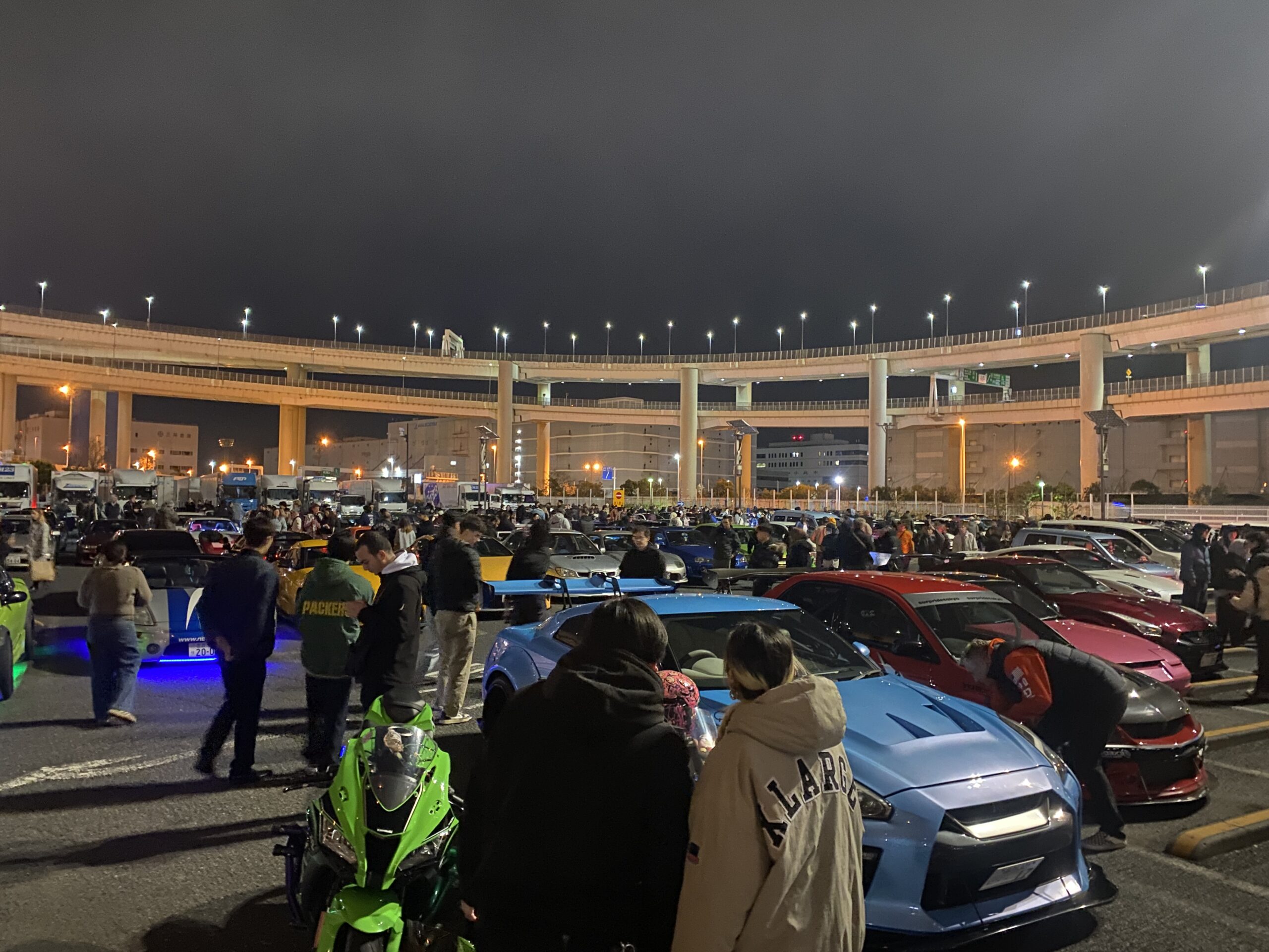 A crowded Daikoku PA car meet in Yokohama at night, featuring a blue Nissan GT-R and other JDM sports cars under the highway lights.